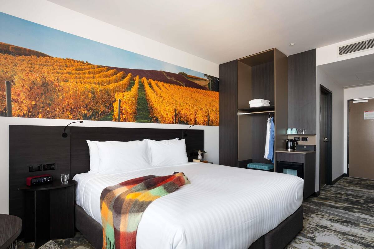 A hotel room with a large bed, a nightstand with a digital clock, a small table with a glass, and a wardrobe with a mirror.