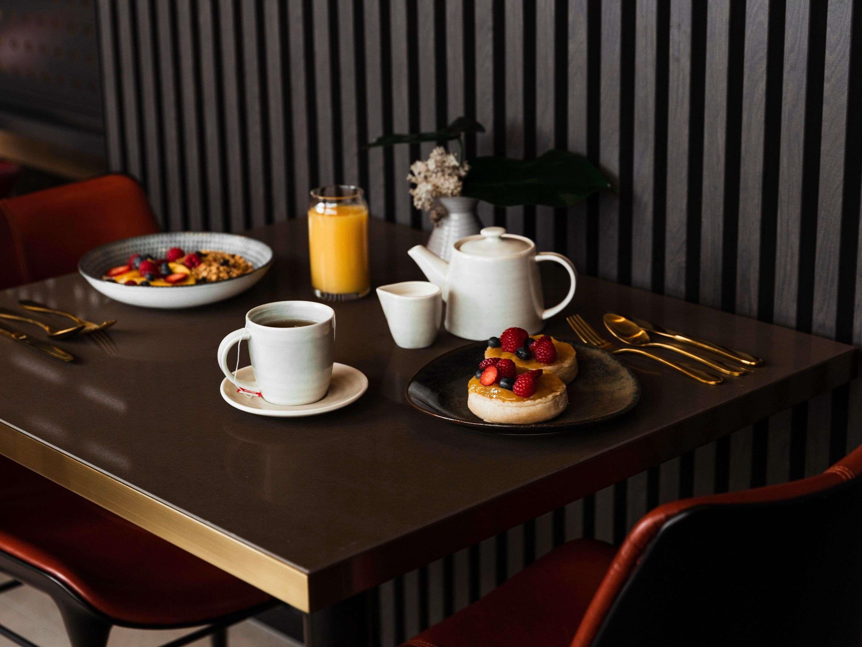 A breakfast table with a bowl of cereal, a cup of coffee, a glass of orange juice, a teapot, and a plate with a dessert topped with berries.