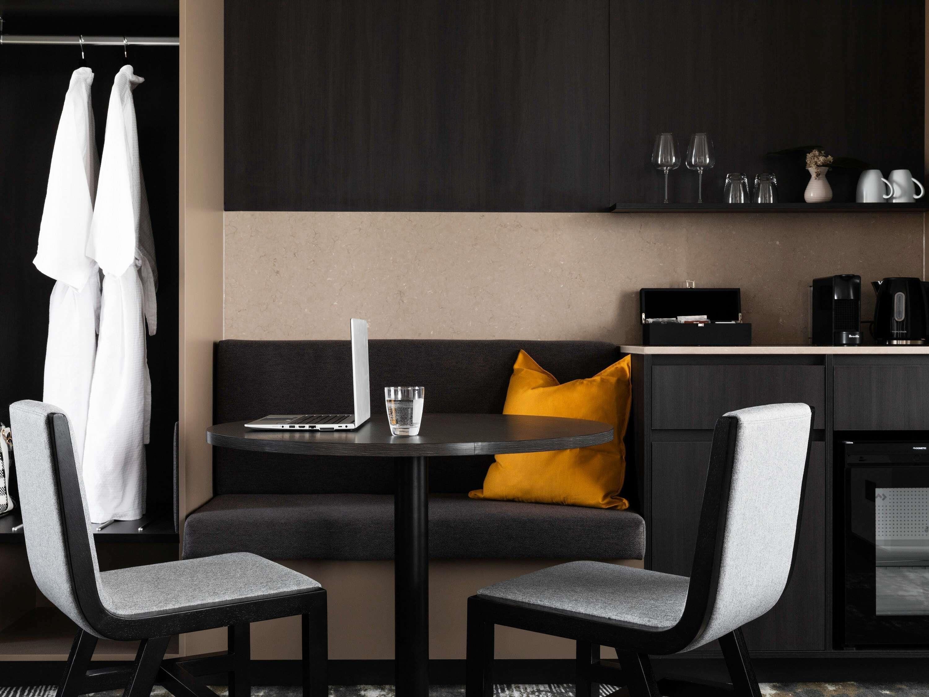 Een moderne hotelkamer met een donkere bank, twee grijze stoelen, een klein rond tafeltje met een laptop en een glas water, en een kitchenette met een koffiezetapparaat en wijnglazen.