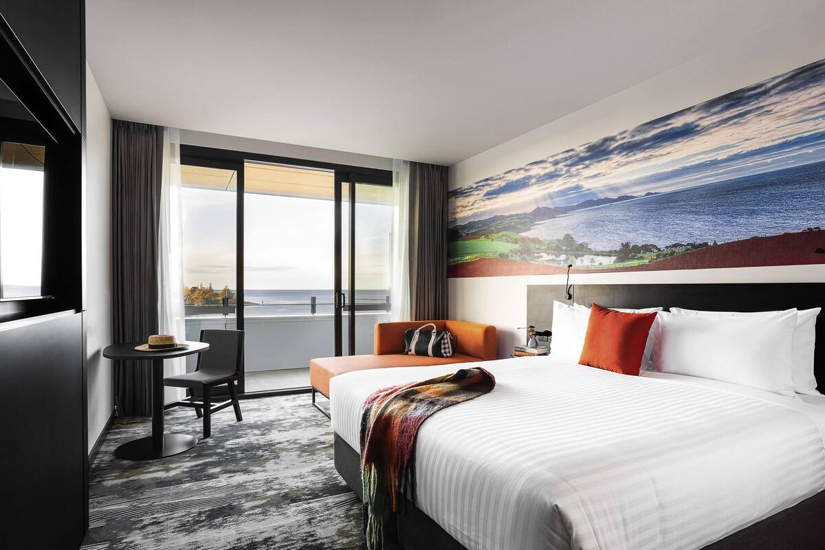 A hotel room with a large bed, a desk, a chair, and a view of the sea through the window.
