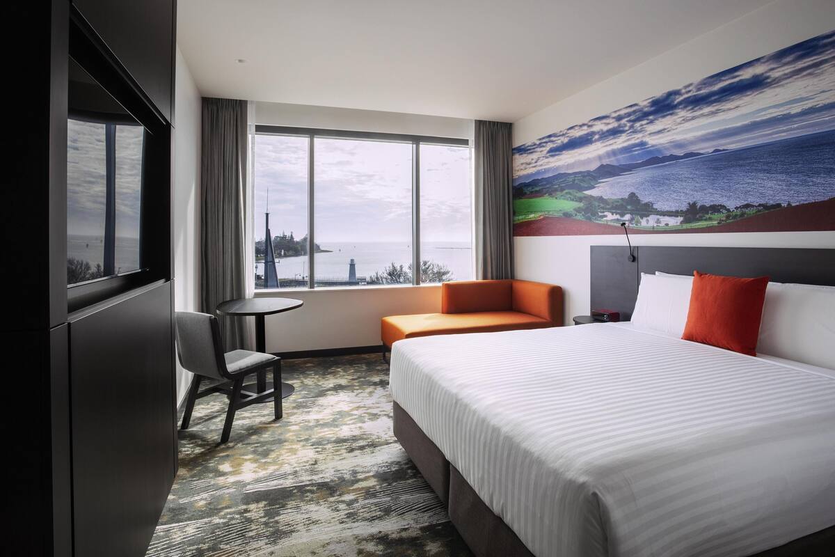A hotel room with a bed, a chair, a small table, and a large window offering a view of the sea and a distant cityscape.