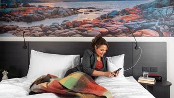 Una persona sentada en una cama con una manta, usando un teléfono inteligente, en una habitación con un cuadro grande de un paisaje rocoso y una mesita de noche con un libro y un vaso.