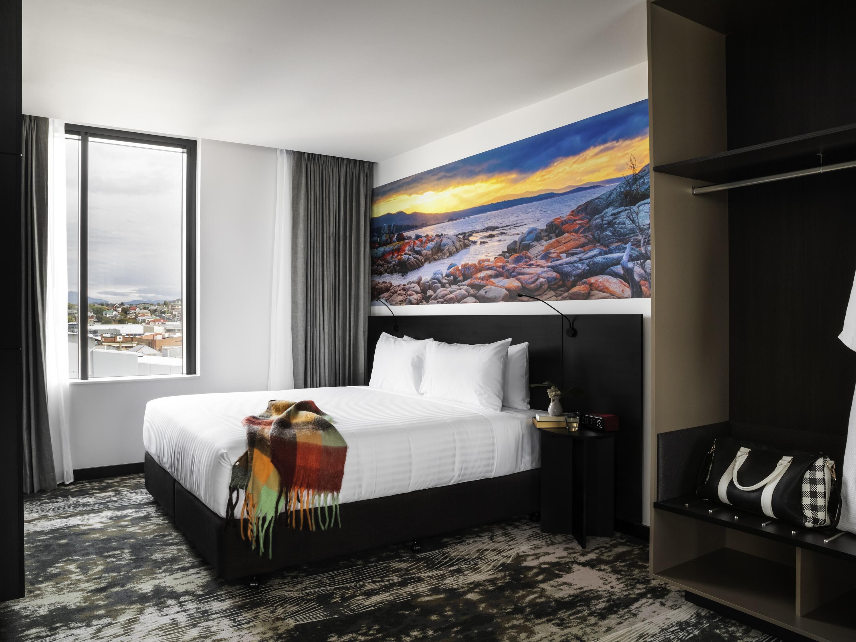 Habitación de hotel con cama, mesita de noche, armario, un cuadro y vistas a la ciudad.