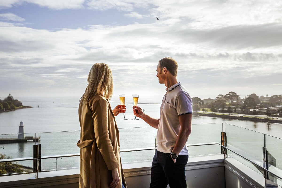 A couple toasting with champagne glasses overlooking a body of water with a distant shoreline.