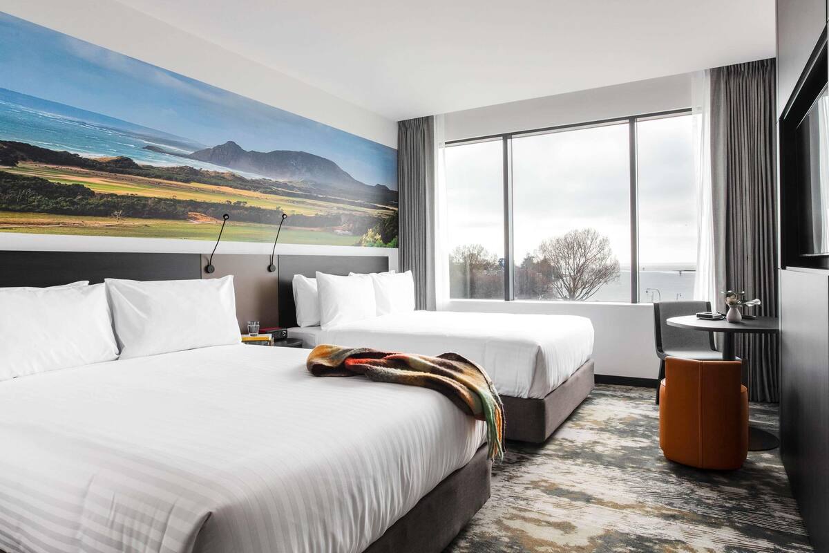 A hotel room with two beds, a large window with a view, a small table, and a chair.