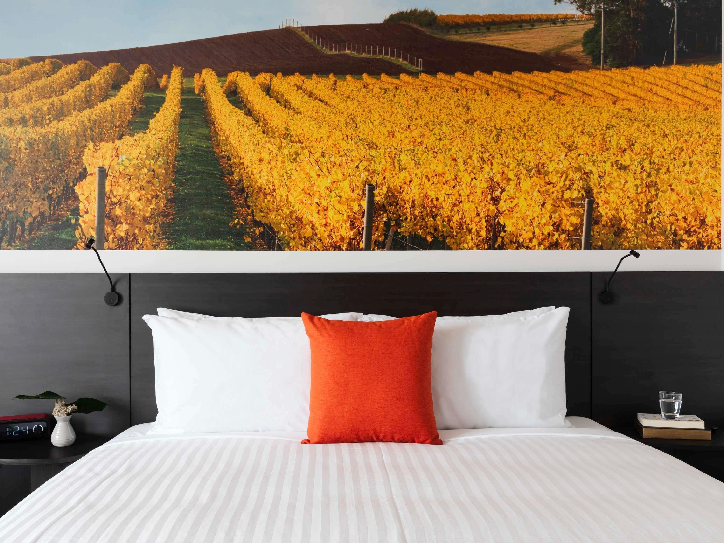 Een hotelkamer met een groot bed, witte beddengoed en een rode kussen. Een digitale klok en een vaas met bloemen staan op het nachtkastje.