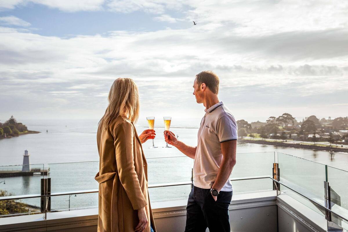 A couple toasting with champagne glasses overlooking a body of water with a distant town and lighthouse.