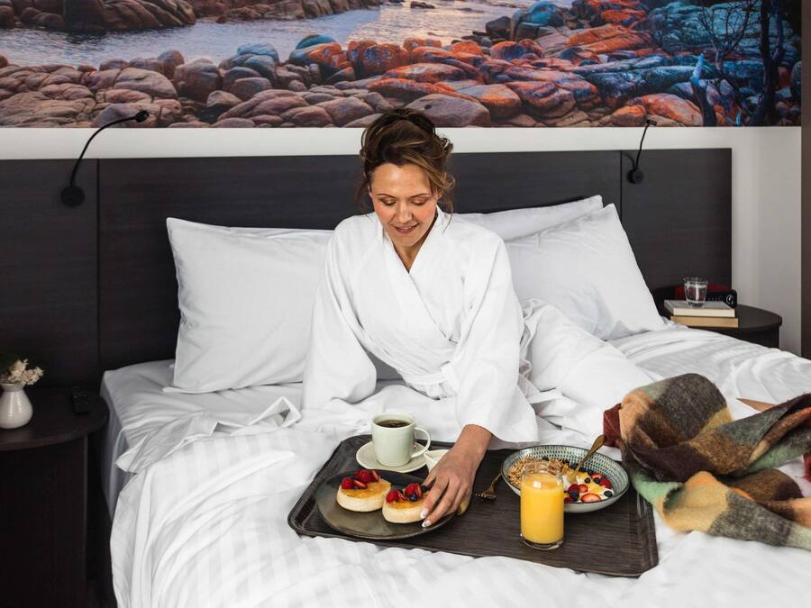 A person in a white bathrobe sitting on a bed with breakfast items, including pancakes, fruit, and orange juice.