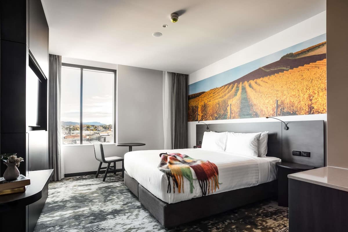 A hotel room with a large bed, a desk, a chair, a view of a cityscape, and a painting of a vineyard.