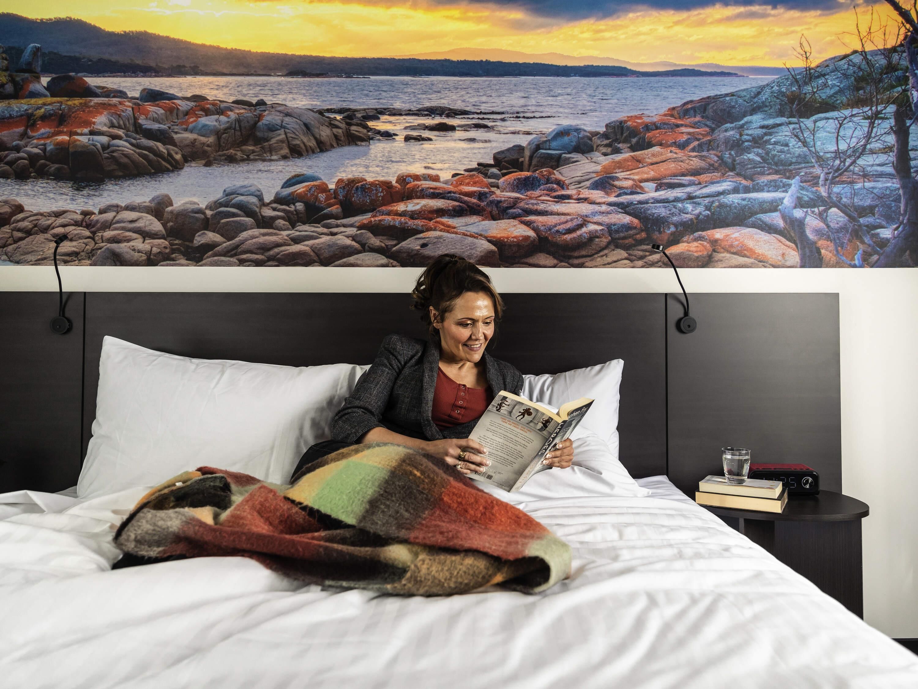 Una persona leyendo un libro en una cama con una manta, una mesita de noche con un libro y un vaso, y una pared con un cuadro de una costa rocosa.