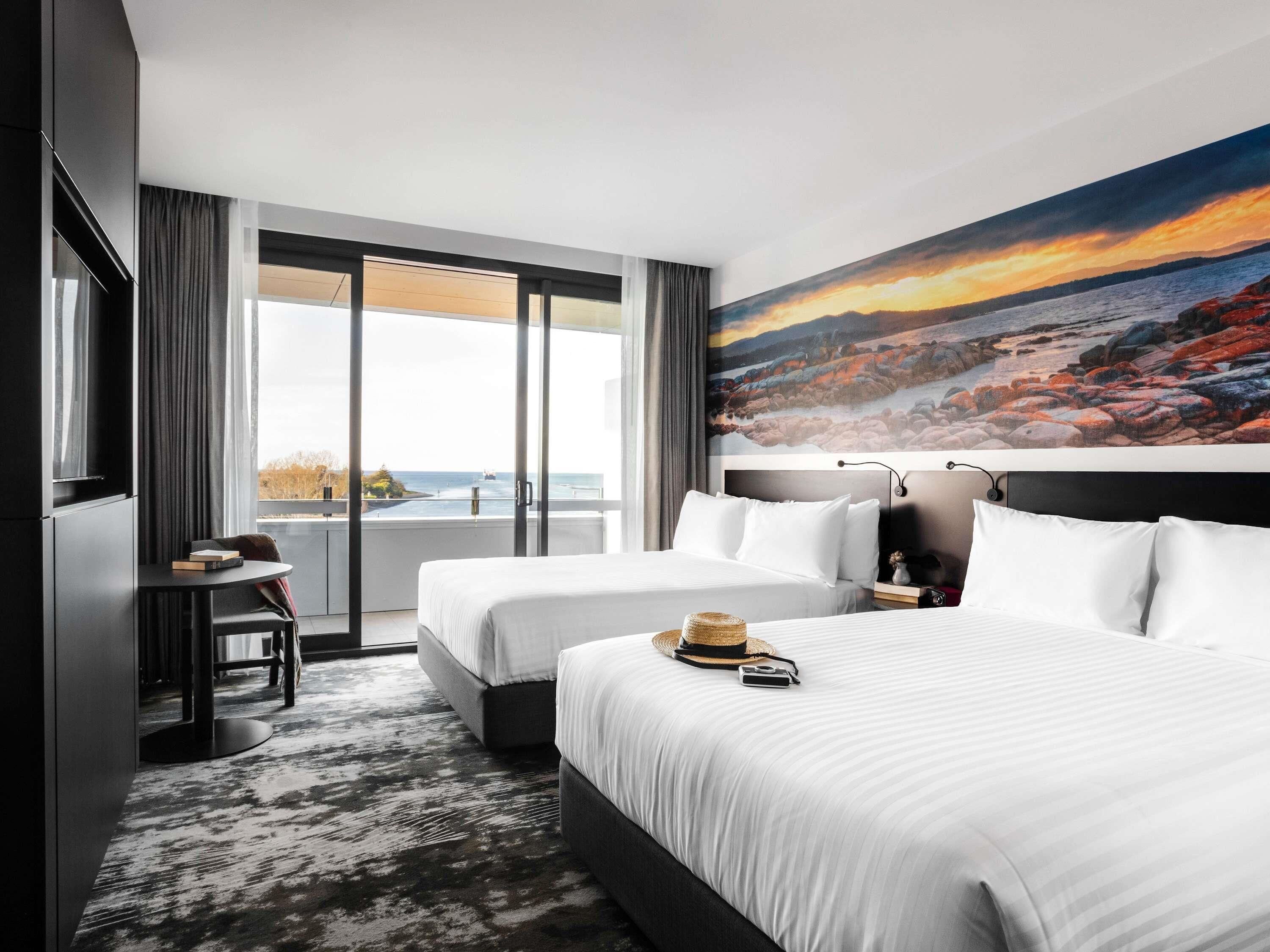 Habitación de hotel con dos camas, un escritorio, una silla, un ventanal con vistas al mar y un cuadro en la pared.