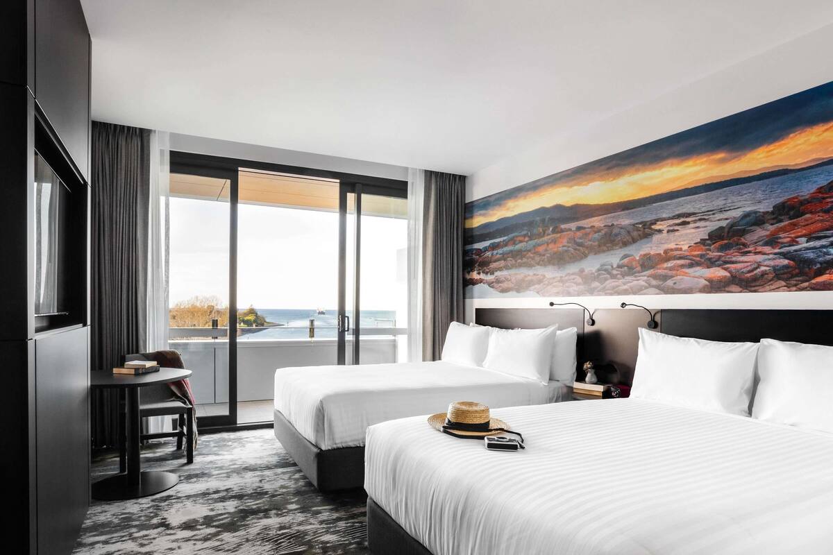 A hotel room with two beds, a desk, a chair, a large window with a view of the sea, and a painting on the wall.