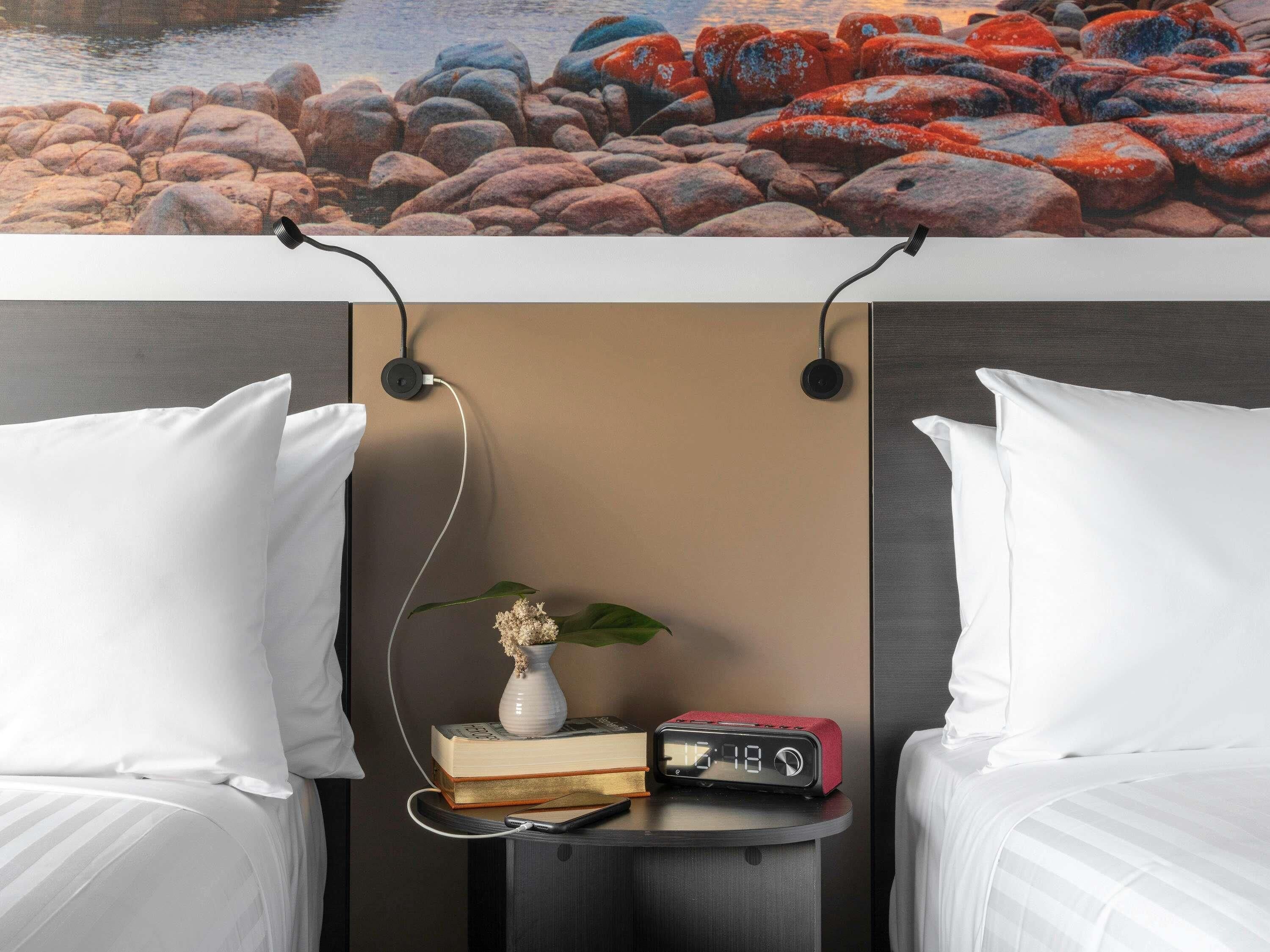 Une chambre d’hôtel avec deux lits, une table de chevet avec une horloge, un vase de fleurs et une vue sur des rochers.
