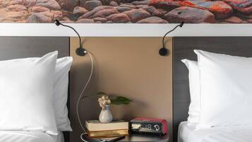 A hotel room with two beds, a bedside table with a clock, a vase with flowers, and a view of rocks.