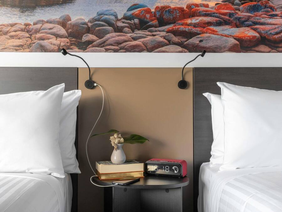 A hotel room with two beds, a bedside table with a clock, a vase with flowers, and a view of rocks.