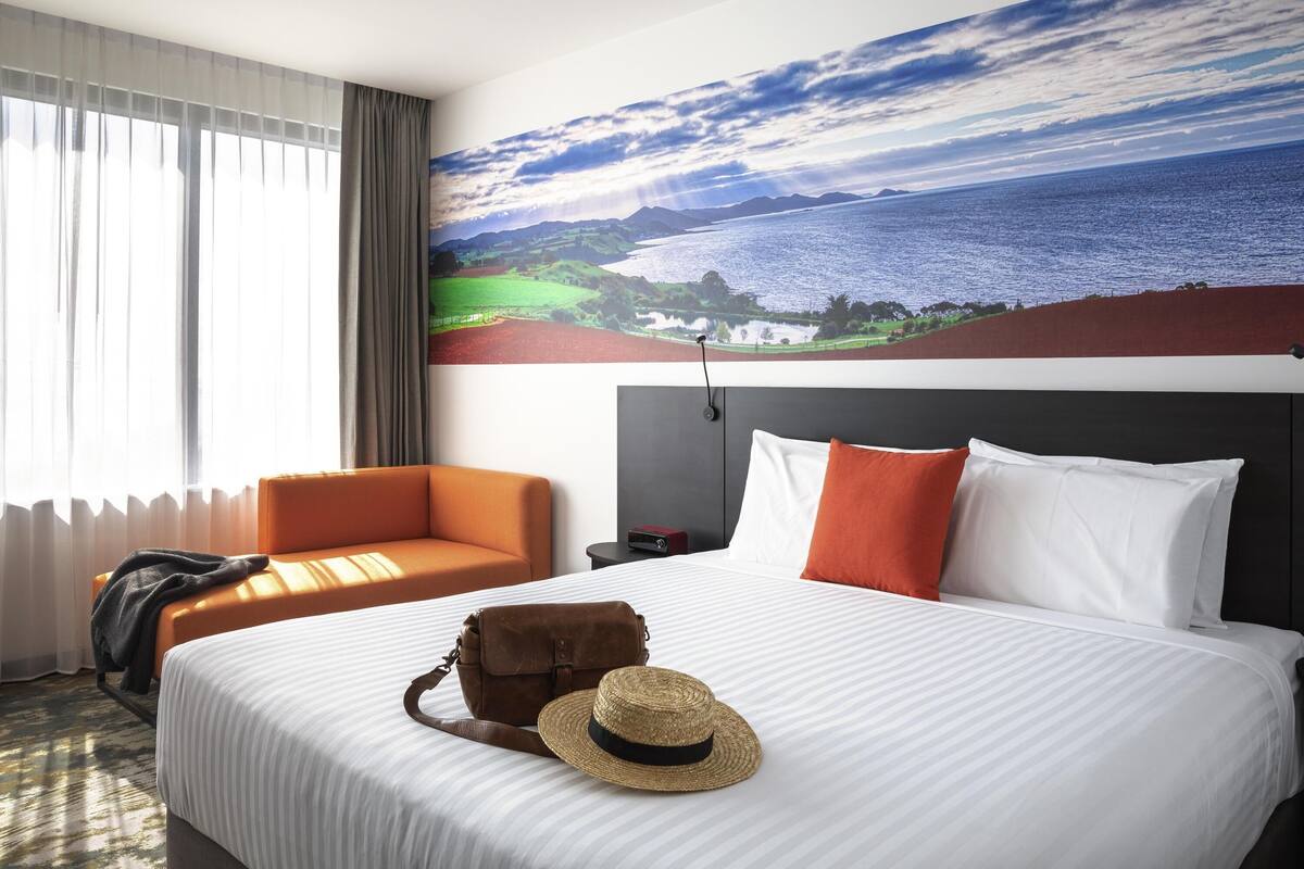 A hotel room with a bed, a chair, a nightstand, a lamp, a painting of a landscape, and a straw hat on the bed.