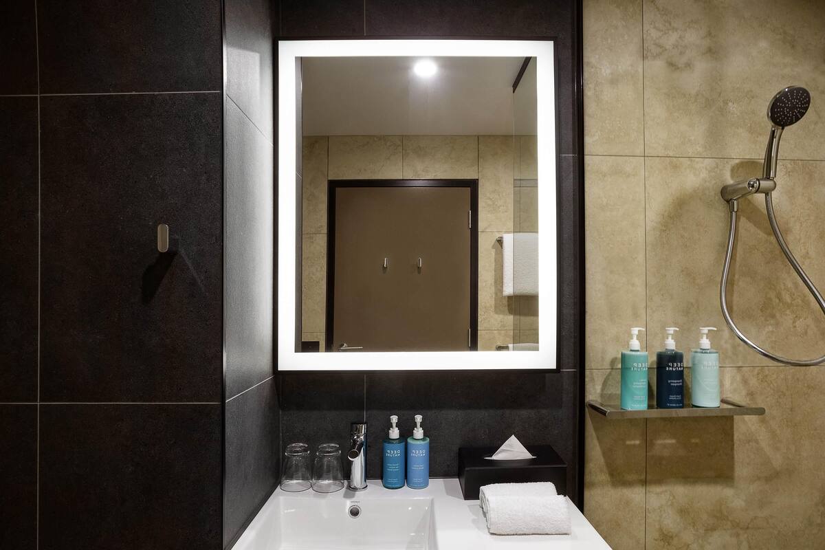 Un baño con un espejo grande, un lavamanos y una ducha. Hay artículos de tocador en el lavamanos y una toalla en la pared.
