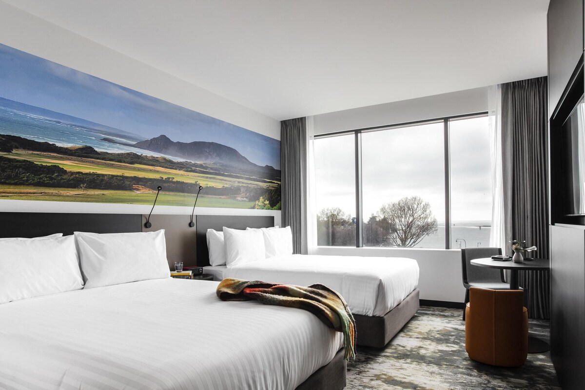A hotel room with two beds, a large window, a painting of a landscape, and a small table with a chair.