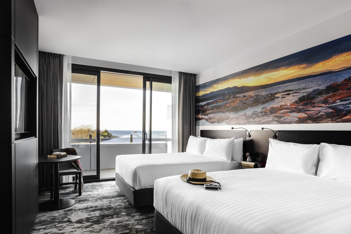 A hotel room with two beds, a desk, a chair, a large window with a view of the sea, and a painting on the wall.