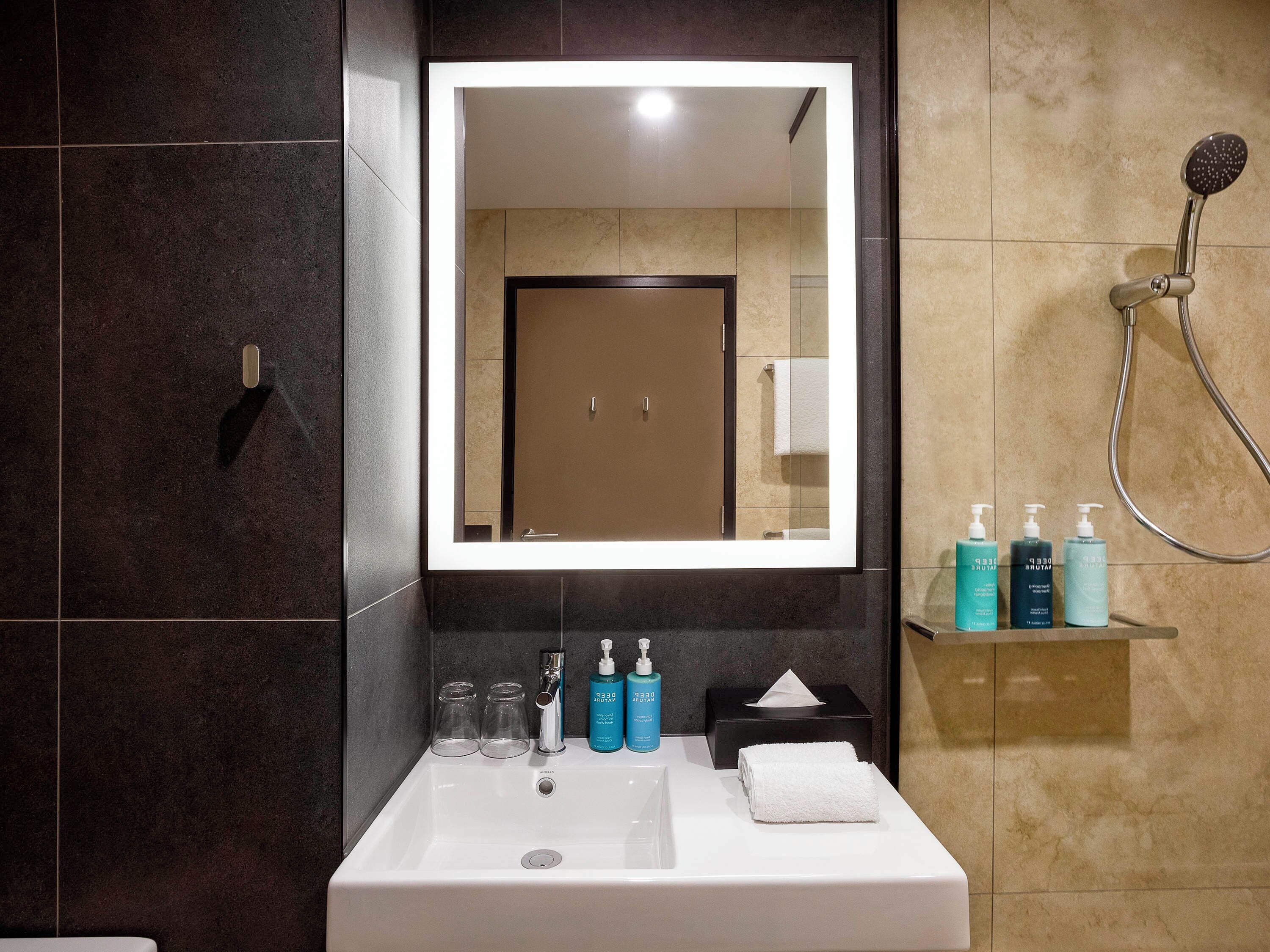 Un baño con un espejo grande, un lavamanos y una ducha. Hay artículos de tocador en el lavamanos y una toalla en la pared.