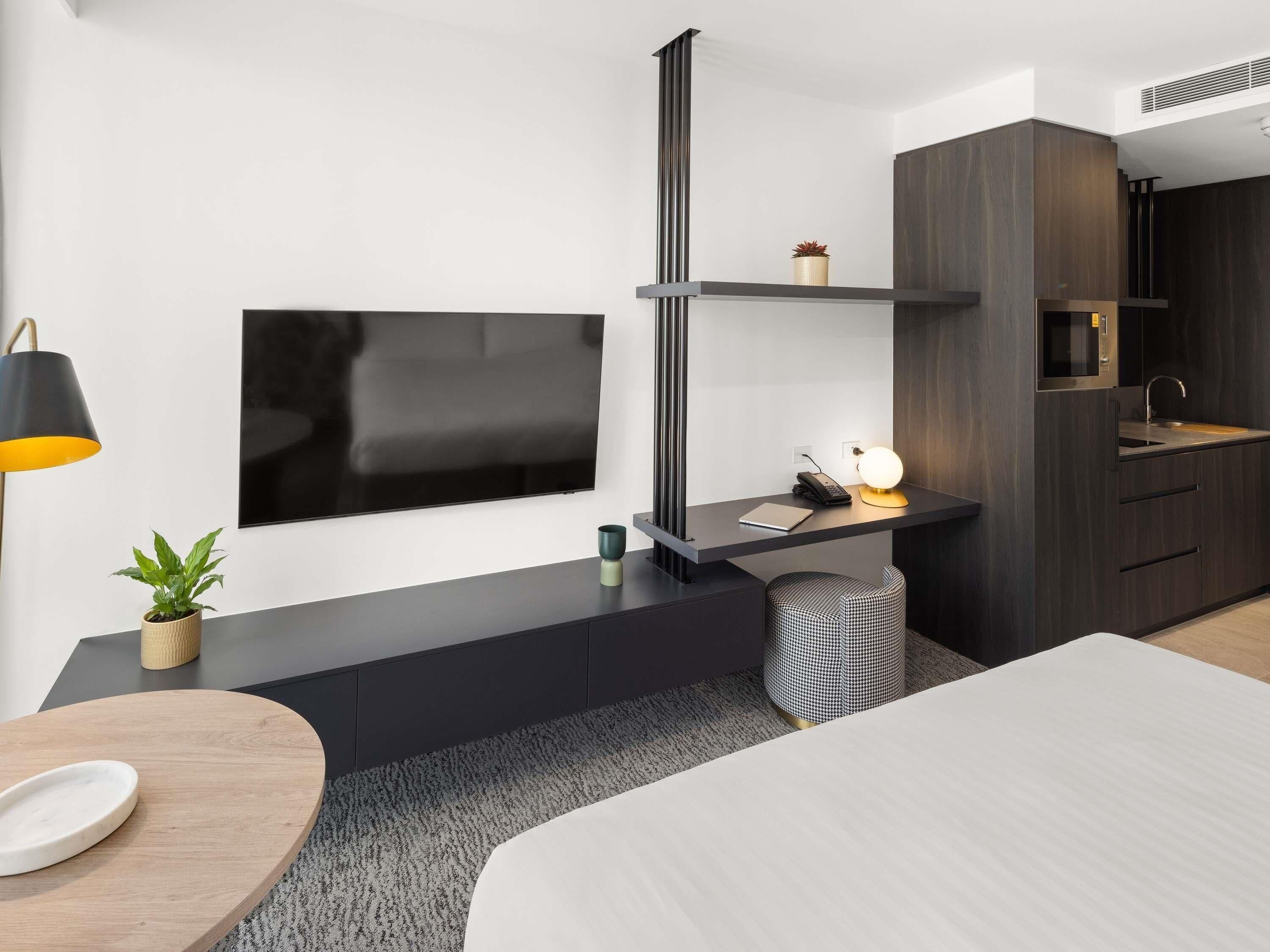 A modern bedroom with a bed, a desk with a lamp, a TV, and a kitchenette.