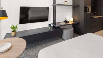 A modern bedroom with a bed, a desk with a lamp, a TV, and a kitchenette.