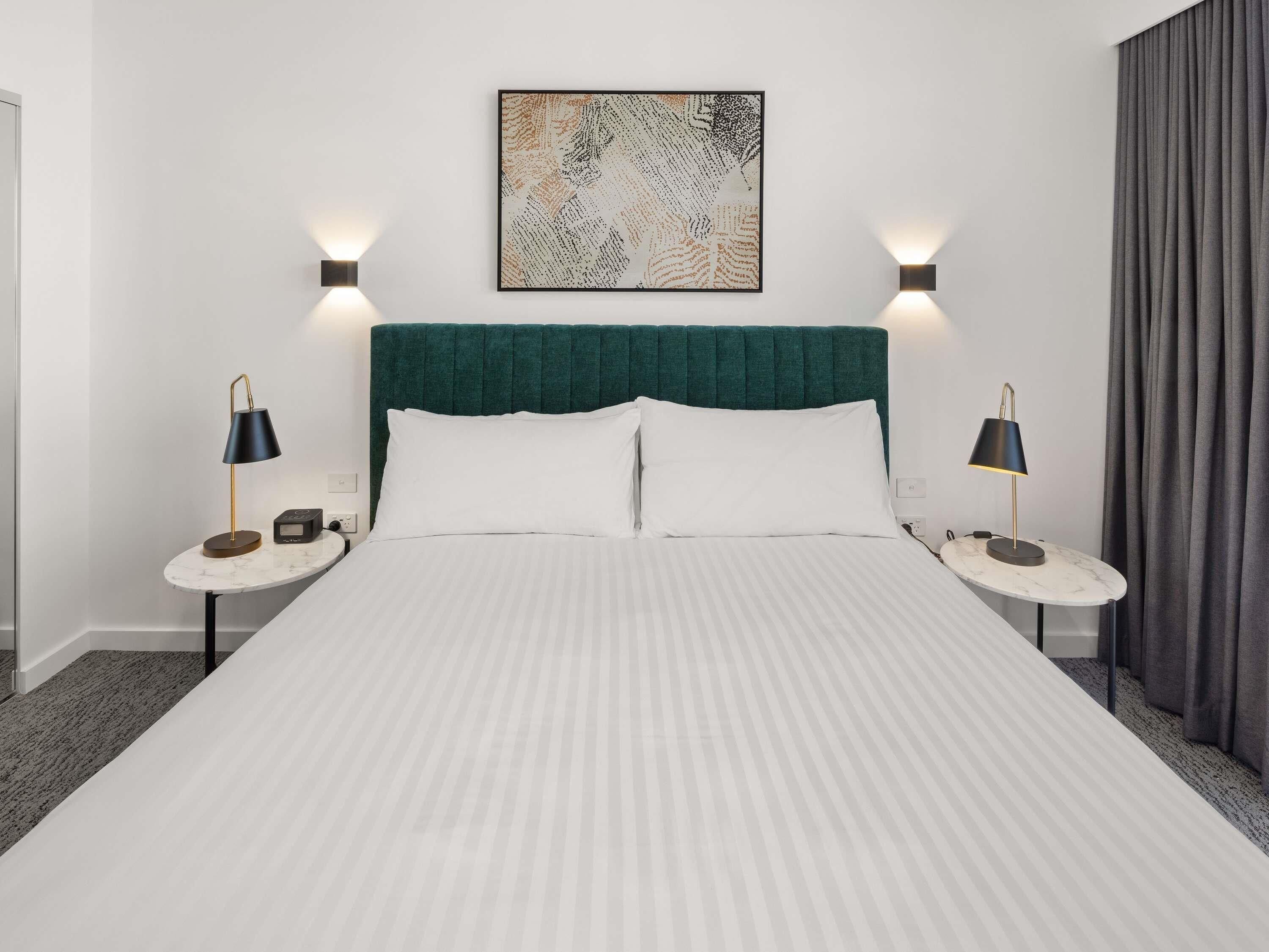 A hotel room with a bed, two bedside tables, a headboard, a wall-mounted artwork, and a window with curtains.