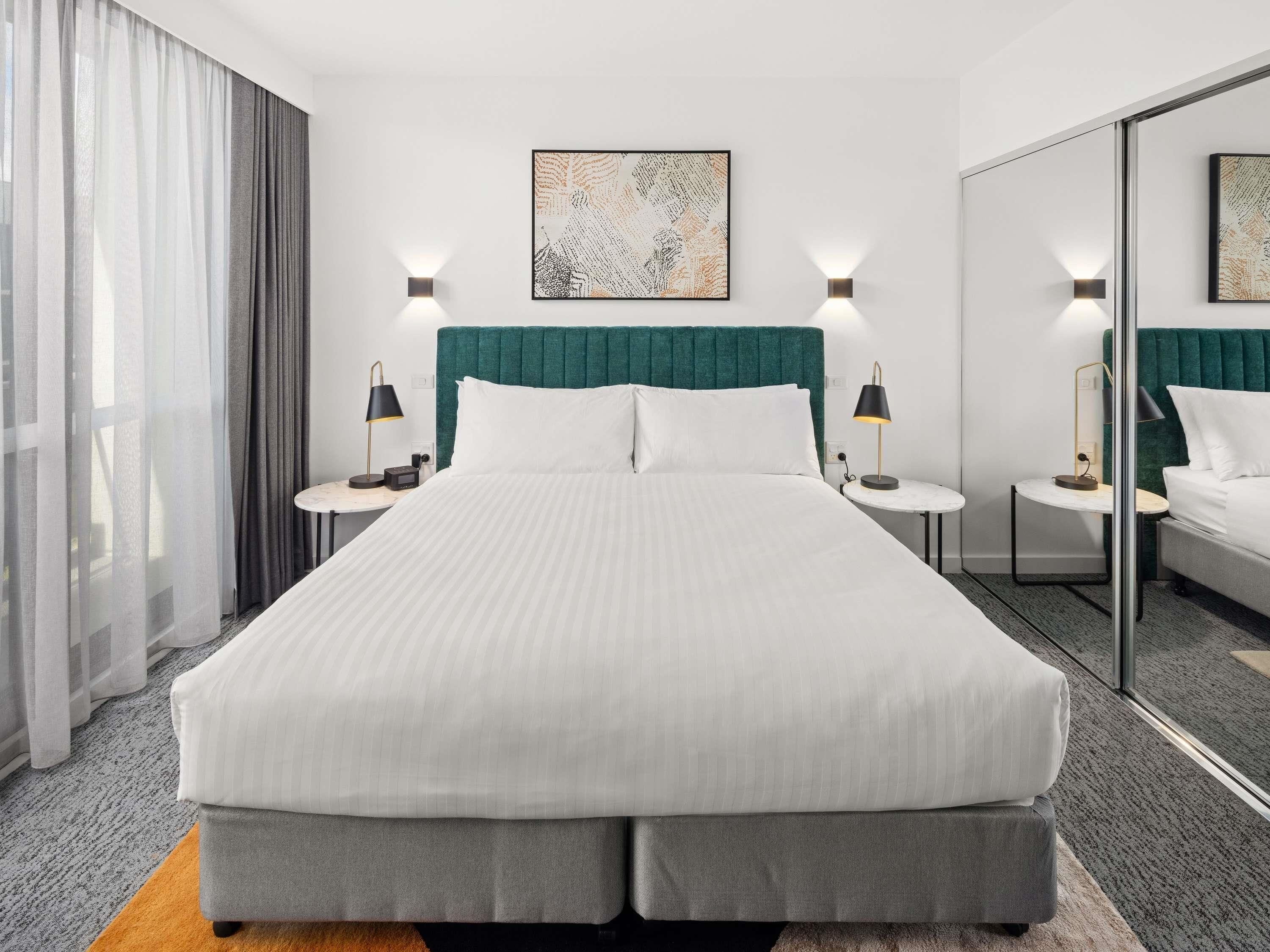 Une chambre d’hôtel avec un grand lit, deux tables de chevet avec des lampes, un miroir et une fenêtre avec des rideaux légers.