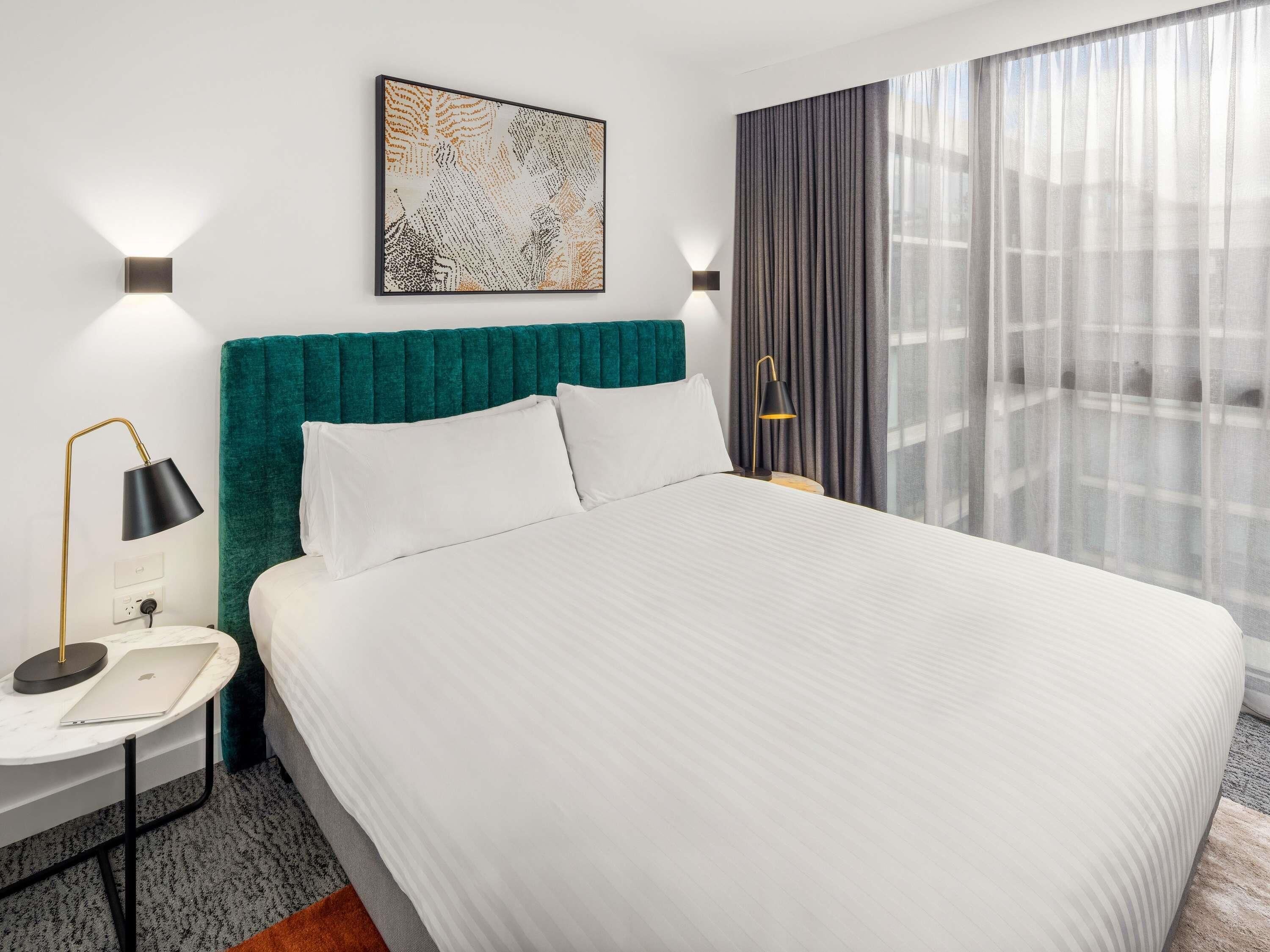 A hotel room with a bed, a desk with a laptop, a painting on the wall, and a window with curtains.