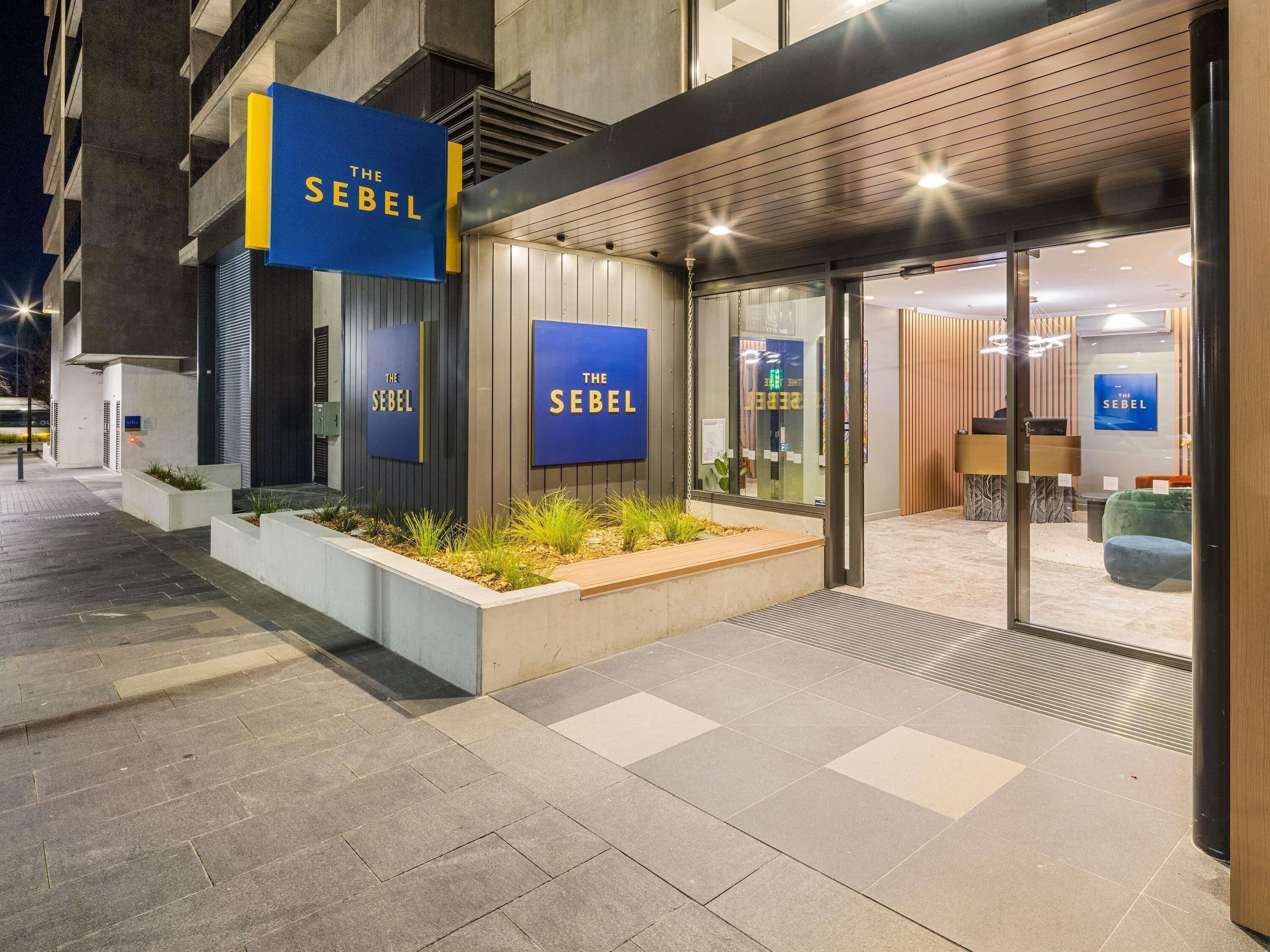 Exterior de un edificio moderno con la señalización 'The Sebel', un área de recepción y una entrada ajardinada.