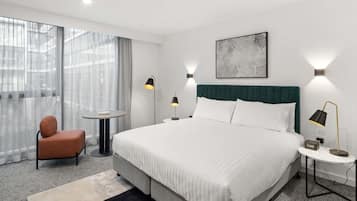 Une chambre d’hôtel moderne avec un grand lit, une chaise, une petite table et une vue sur la ville à travers de fines rideaux.