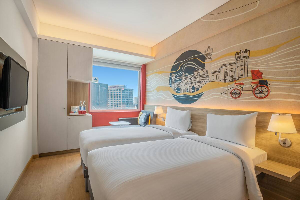 A hotel room with two beds, a TV, a window with a city view, and a mural on the wall.