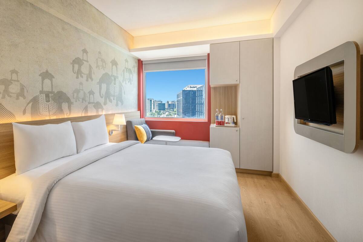 A hotel room with a large bed, a nightstand, a TV mounted on the wall, and a window with a city view.