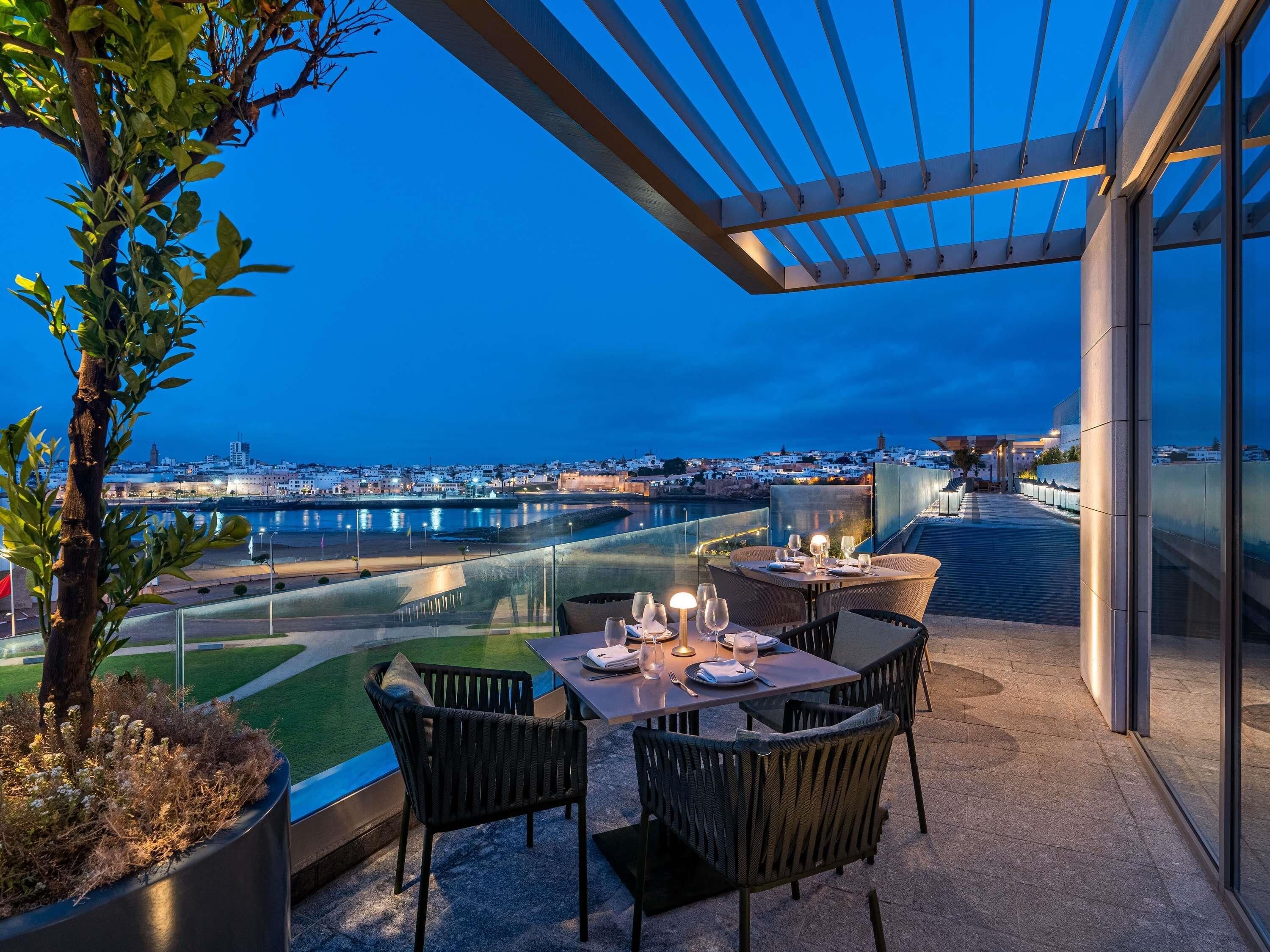 An outdoor dining area with tables and chairs, overlooking a cityscape and waterfront.