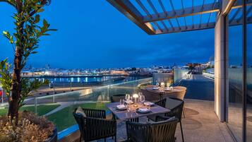 An outdoor dining area with tables and chairs, overlooking a cityscape and waterfront.
