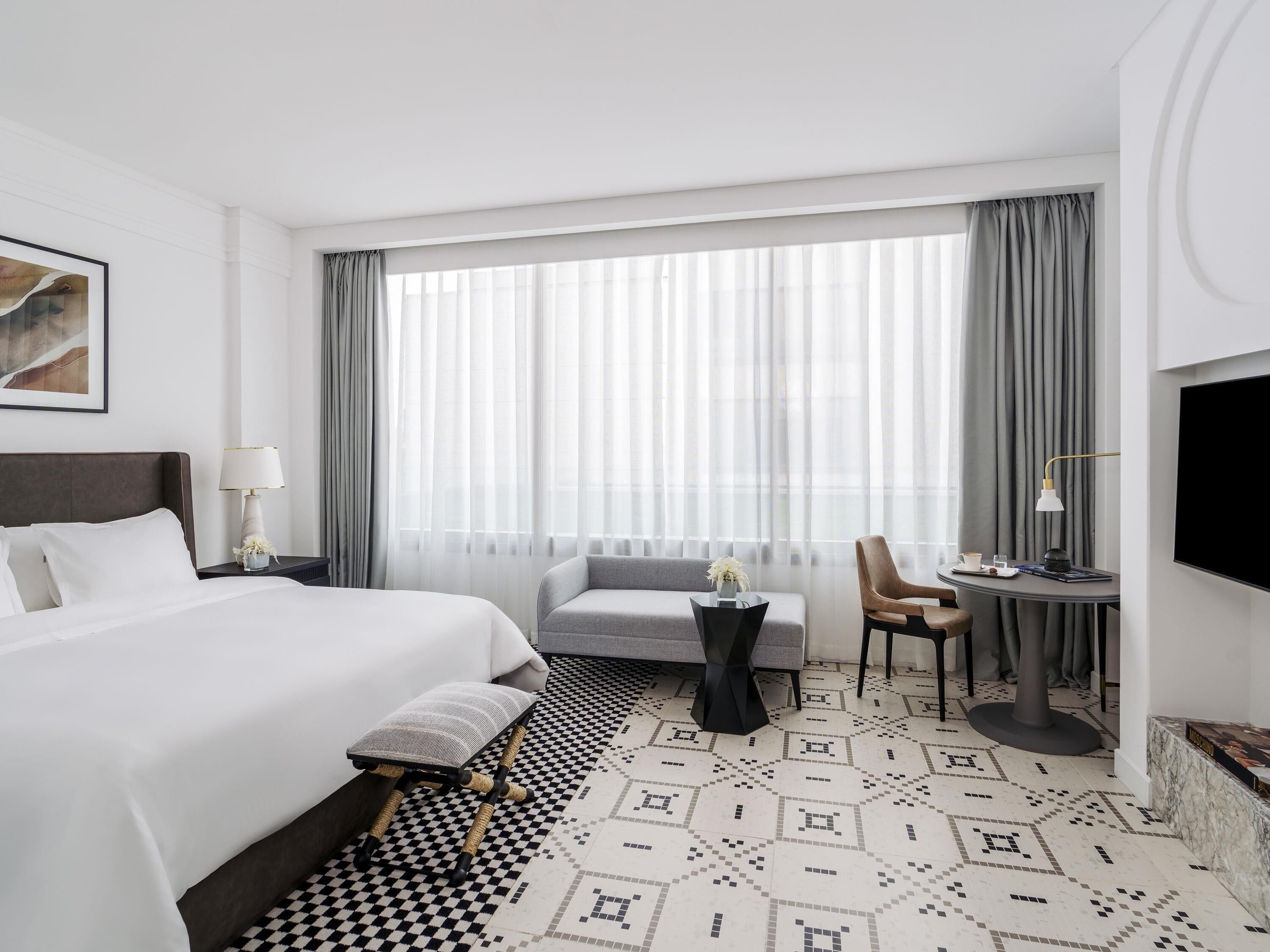 A hotel room with a large bed, a sofa, a small table with a lamp, and a TV mounted on the wall.