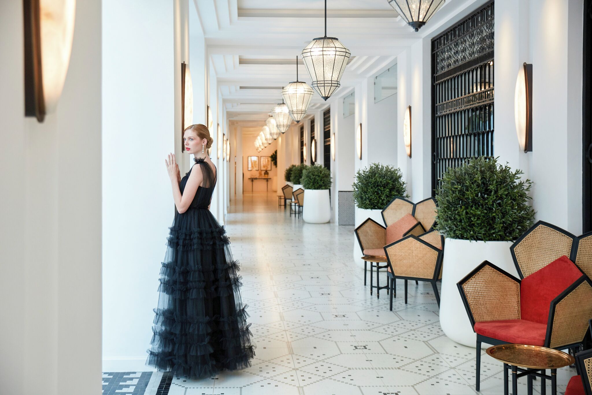 A hotel corridor with a woman in a black dress, hexagonal patterned floor, wicker chairs, and hanging light fixtures.