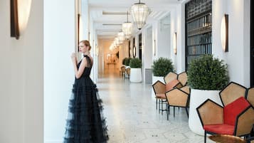A hotel corridor with a woman in a black dress, hexagonal patterned floor, wicker chairs, and hanging light fixtures.