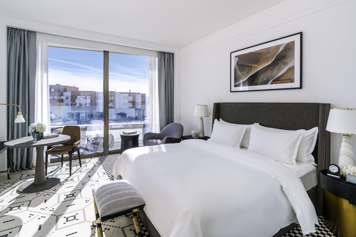 A hotel room with a large bed, a desk with a chair, a small table with a lamp, and a view of buildings through large windows.