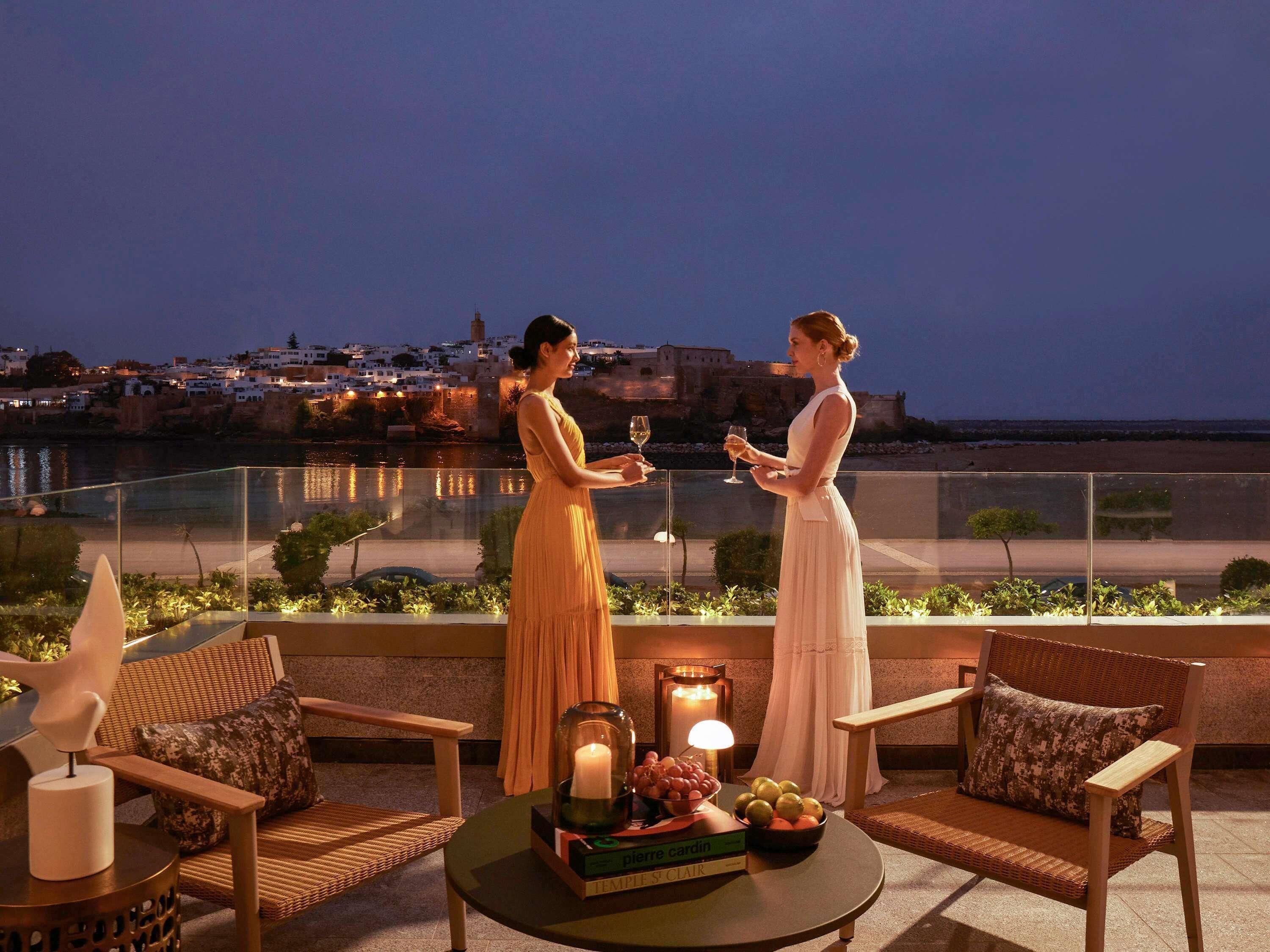 A terrace with two people holding wine glasses, a table with fruit and a candle, and a view of a coastal town at dusk.