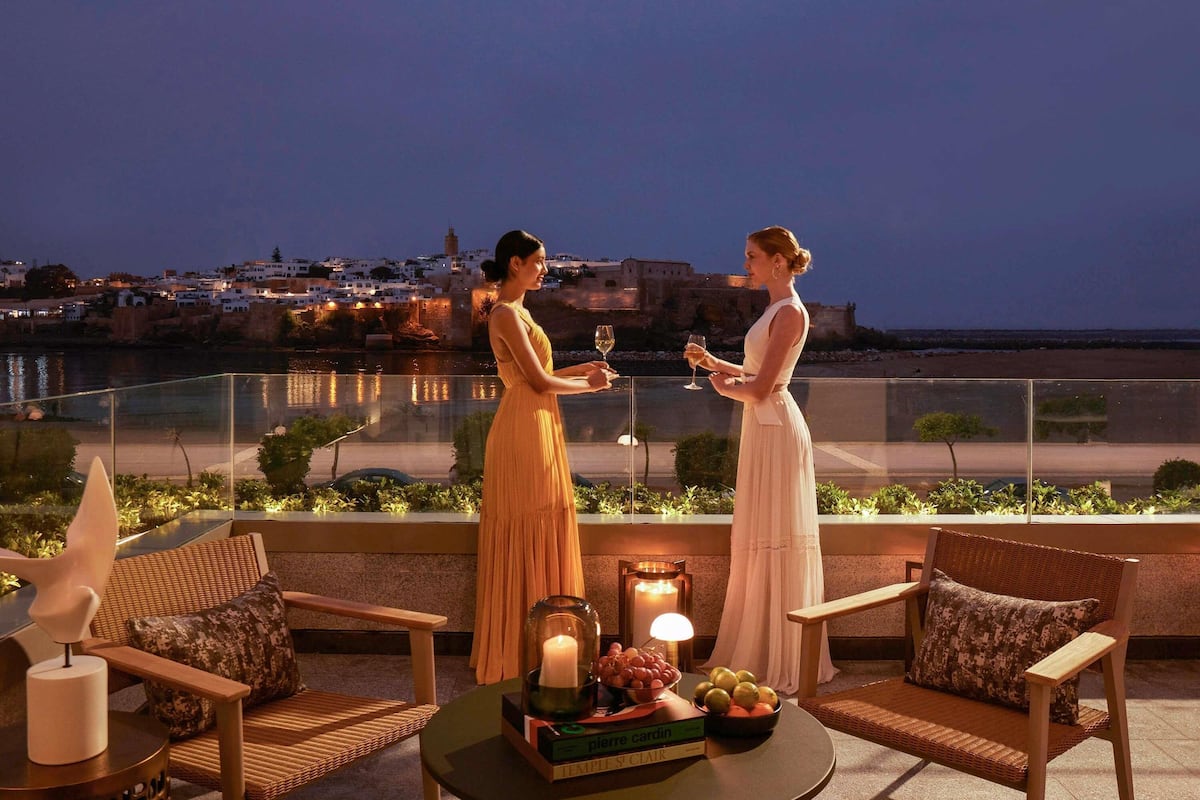 A terrace with two people holding wine glasses, a table with fruit and a candle, and a view of a coastal town at dusk.