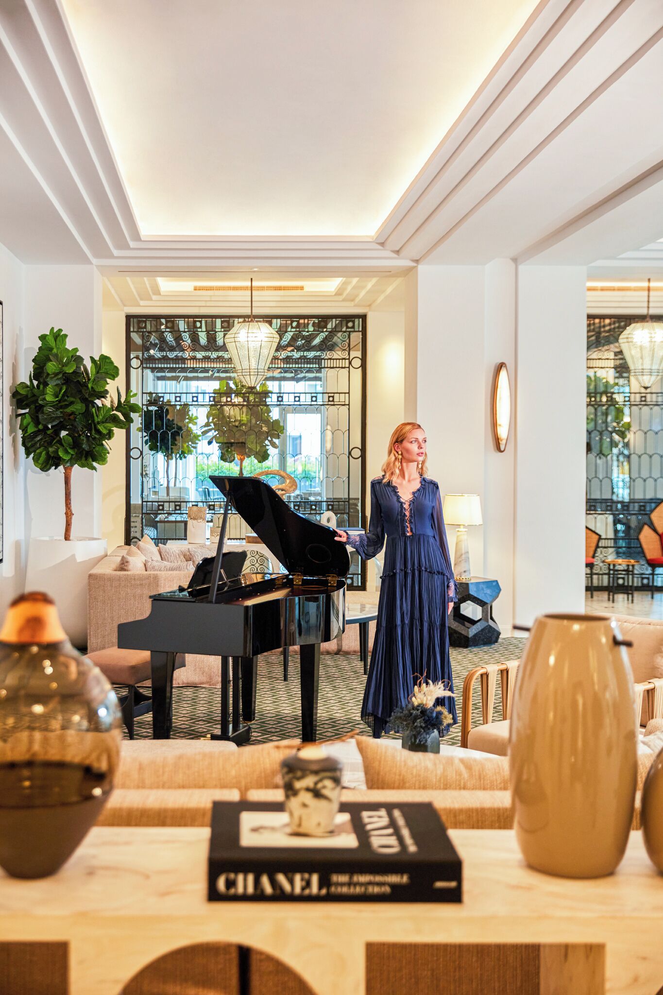 A woman standing near a grand piano in a well-lit, elegant room with a sofa, decorative vases, and a book titled "CHANEL".