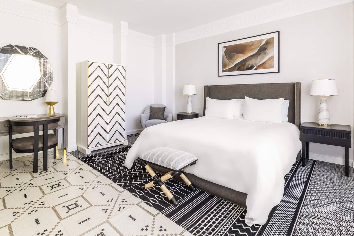 A hotel room with a large bed, a desk with a chair, a side table with a lamp, and a patterned rug.