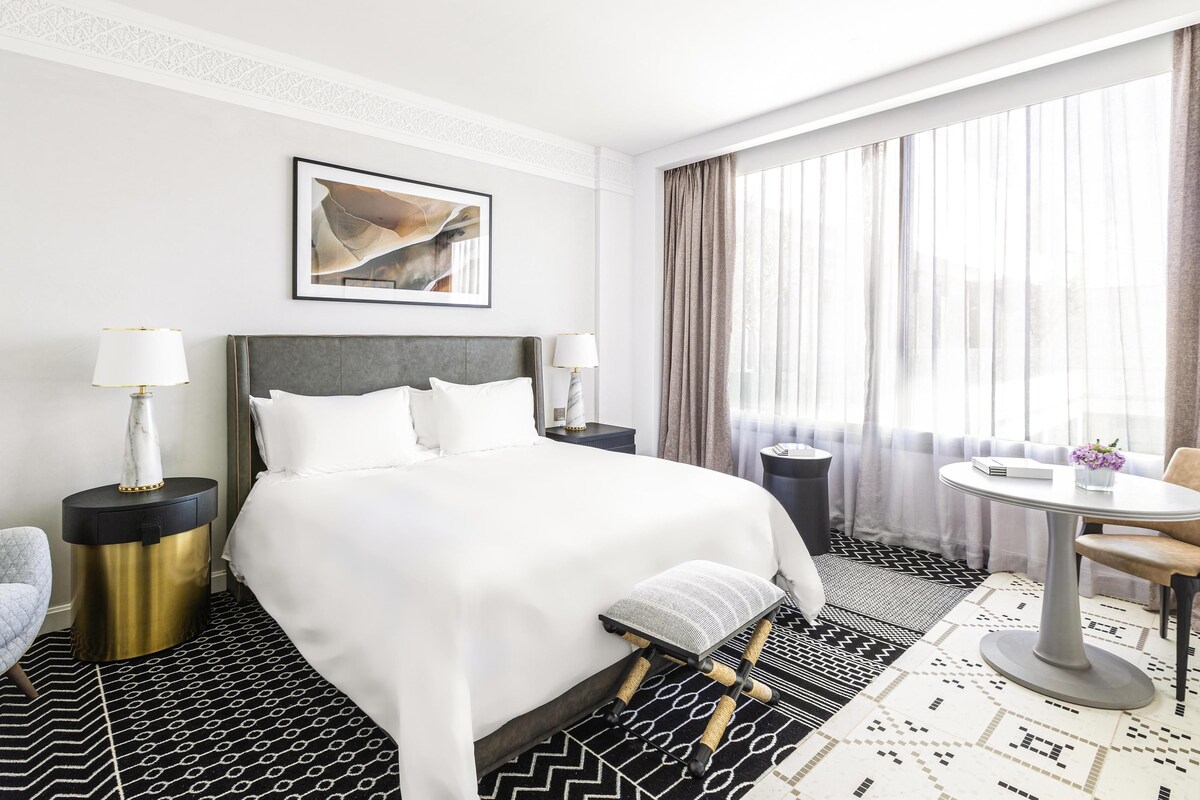 A hotel room with a bed, two bedside tables, a chair, a round table, and a window with curtains.