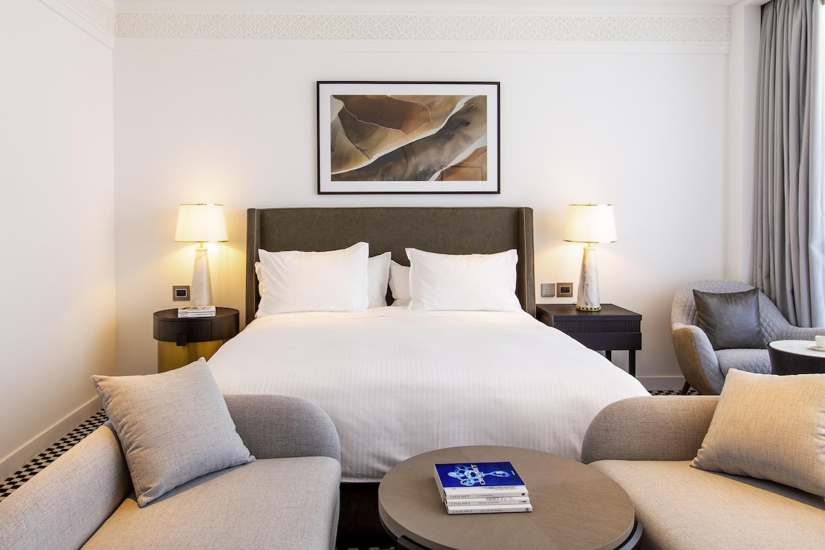 Hotellihuone, jossa on sÀnky, kaksi yöpöytÀlamppua, kehystetty abstrakti taideteos, sohva, pyöreÀ sohvapöytÀ ja tuoli.