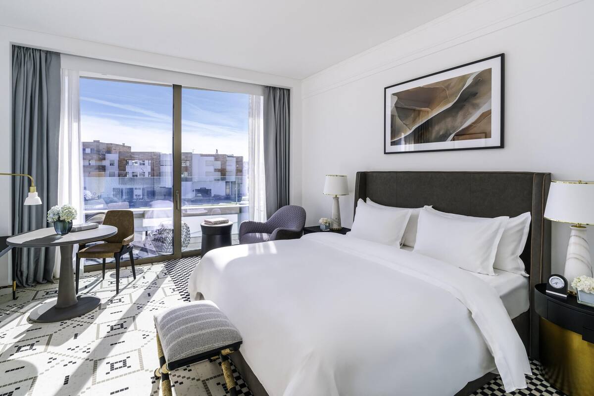 A hotel room with a large bed, a desk with a chair, a small table with a lamp, and a view of buildings through large windows.