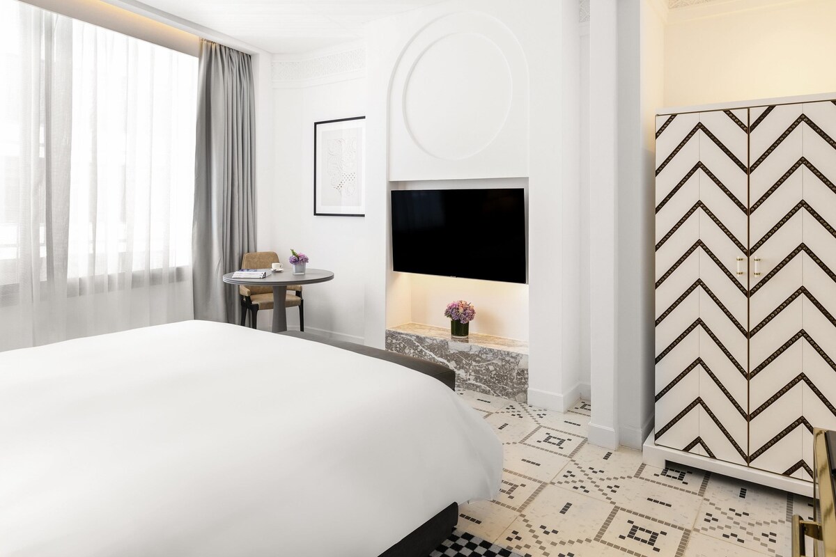 A hotel room with a large bed, a small table with a vase, a flat-screen TV, and a decorative wall panel.