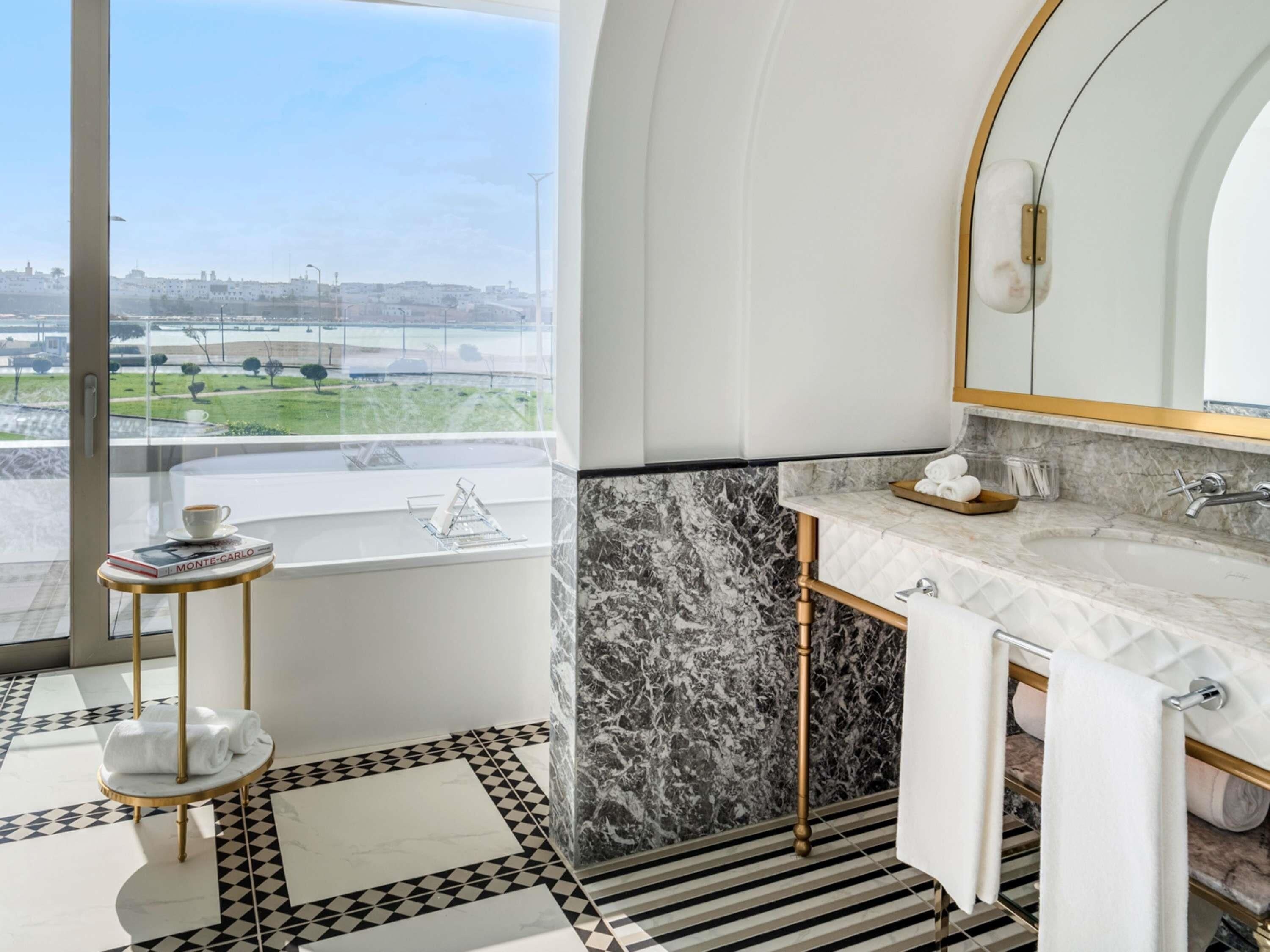 A modern bathroom with a large window, a marble backsplash, a white sink, and a gold-colored towel rack.