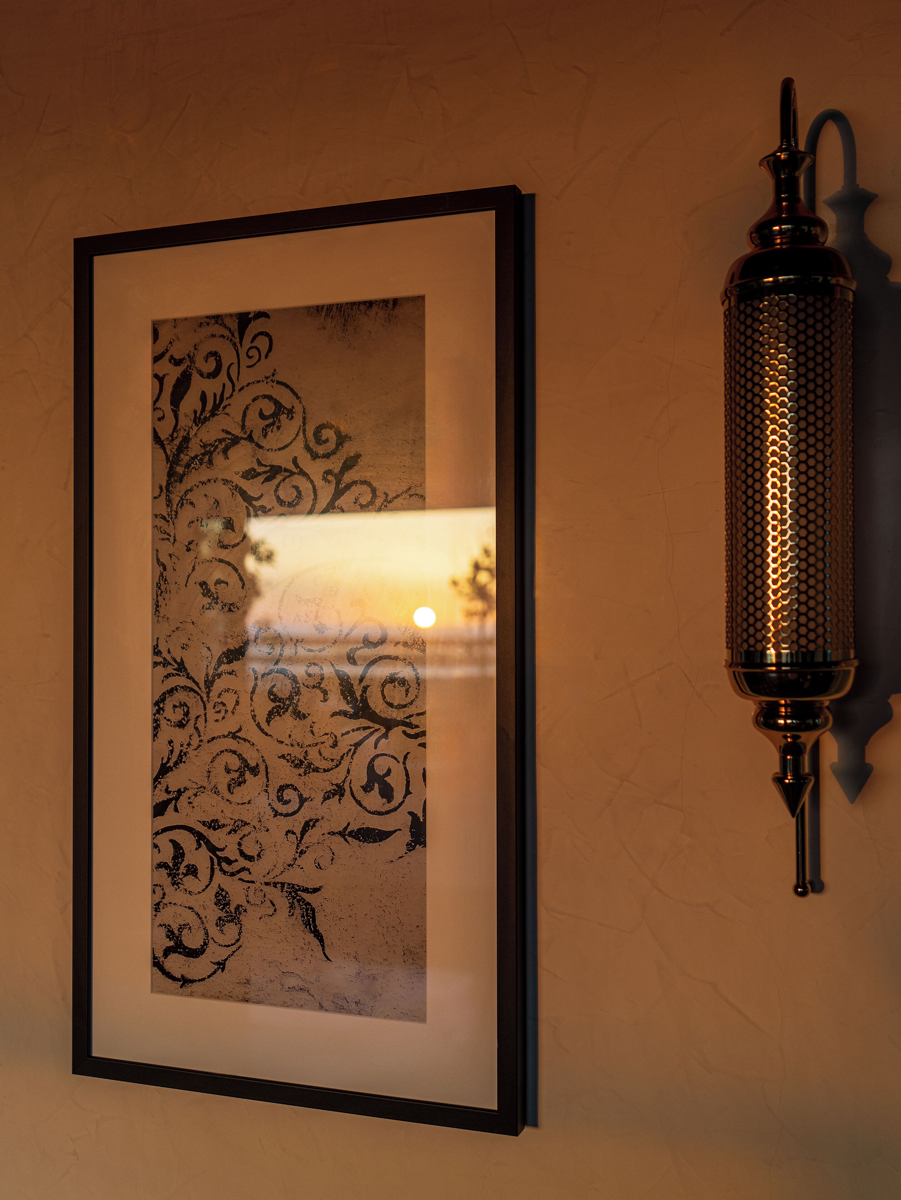 A framed artwork with intricate designs on a wall, next to a wall-mounted, textured light fixture.