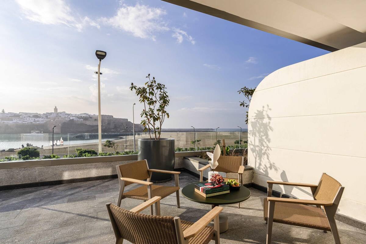A terrace with wicker furniture, a round table with books and fruit, and a view of the sea and a distant cityscape.